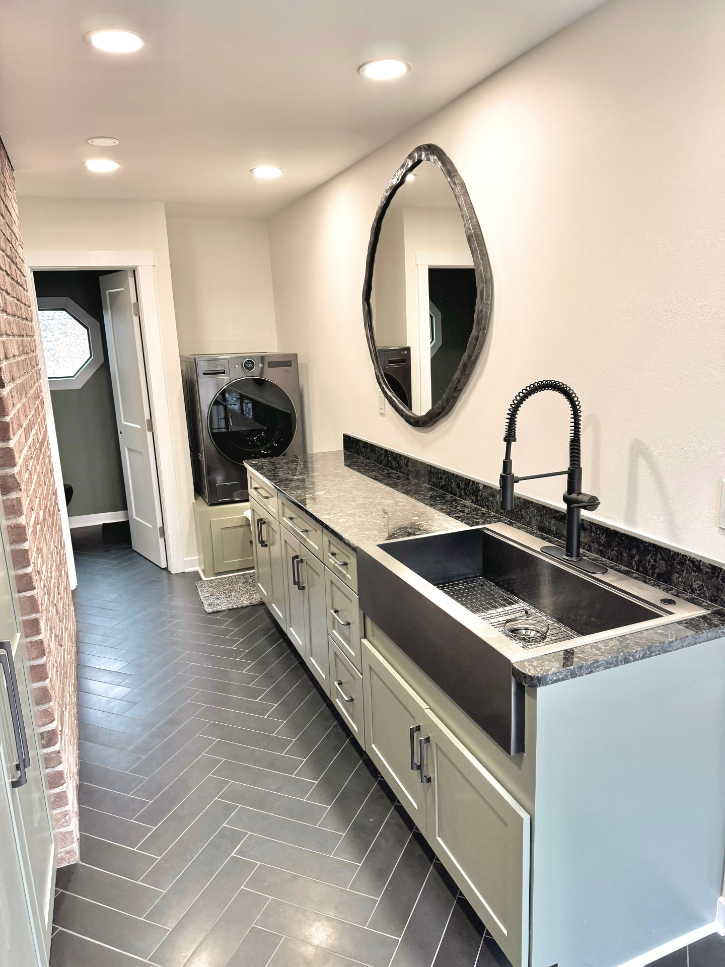 Laundry room remodel with olive green cabinets, dark countertops, and a round mirror